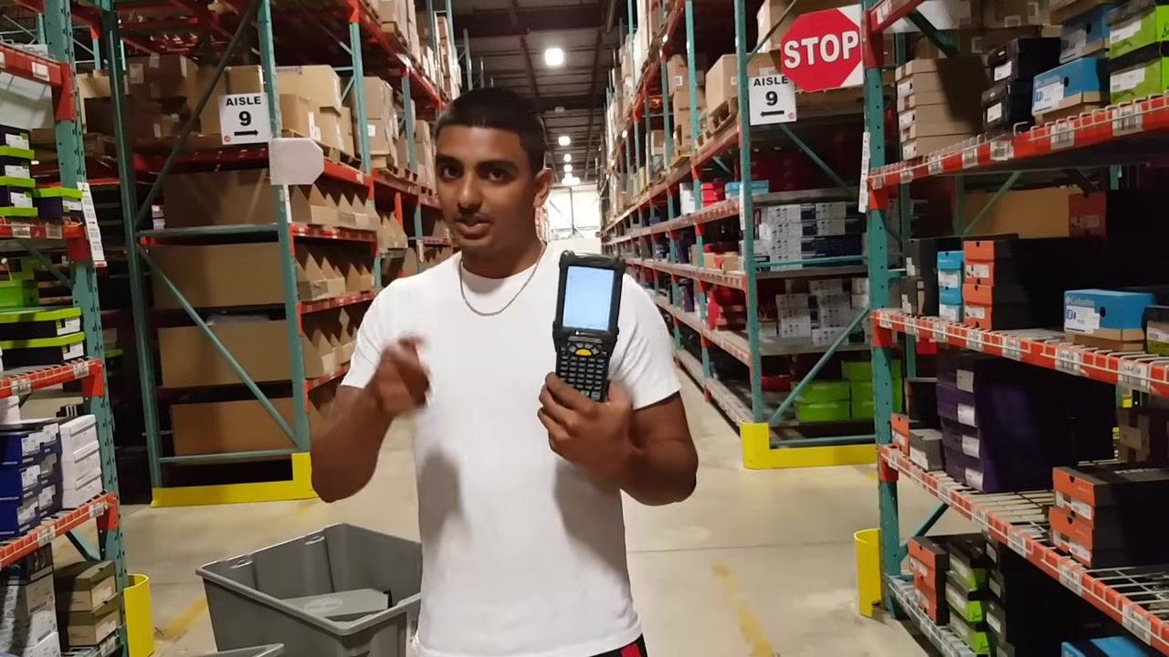 A worker in a white shirt stands in a warehouse aisle, holding a handheld scanner upright and gesturing with his other hand. Shoe boxes line the shelves, and "STOP" and "AISLE 9" signs are visible. Gray totes are on the floor.