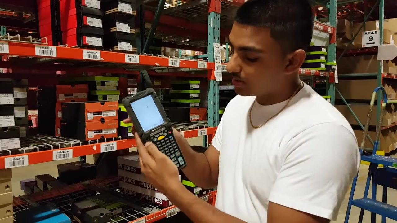 A warehouse worker holds a Motorola RF gun, entering login credentials on the touchscreen. Shoe boxes and shelf labels are visible in the background.
