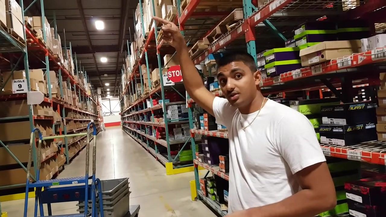 Warehouse worker points upward to high storage locations on top racks, indicating where inbound products are initially stored. Aisle signs, shelves, and boxes are visible.