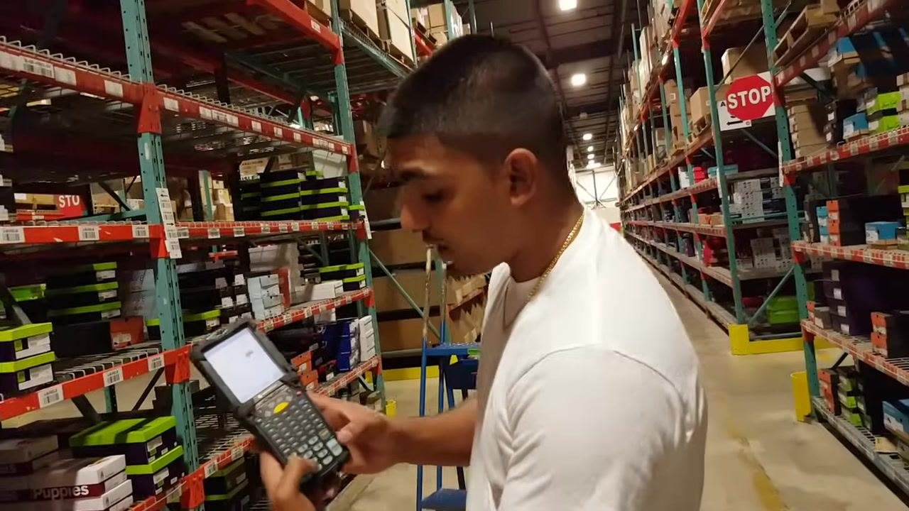 A warehouse worker stands in an aisle, holding a Motorola RF gun and selecting options on the device. Shoe boxes are stacked on shelves, and a blue step ladder and STOP sign are visible in the background.