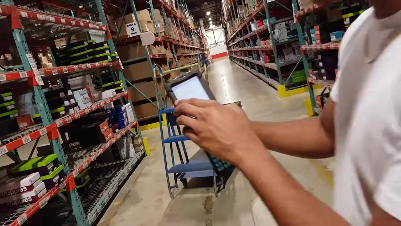 A worker stands in the warehouse aisle, holding the RF gun and preparing to locate the assigned shelf. The Aisle 9 sign and shoe boxes are visible.
