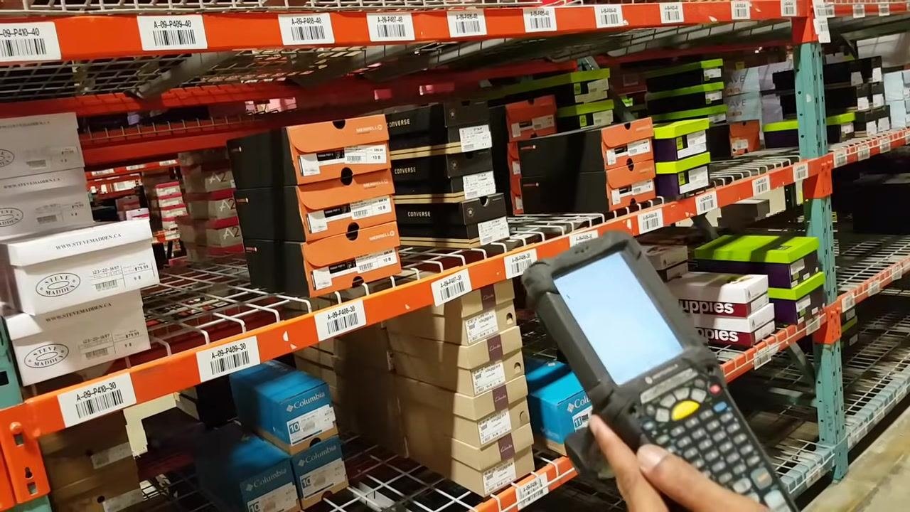 The worker holds the RF gun and points to shelf labels, explaining the meaning of location codes. Shoe boxes and barcode labels are visible on the shelves.