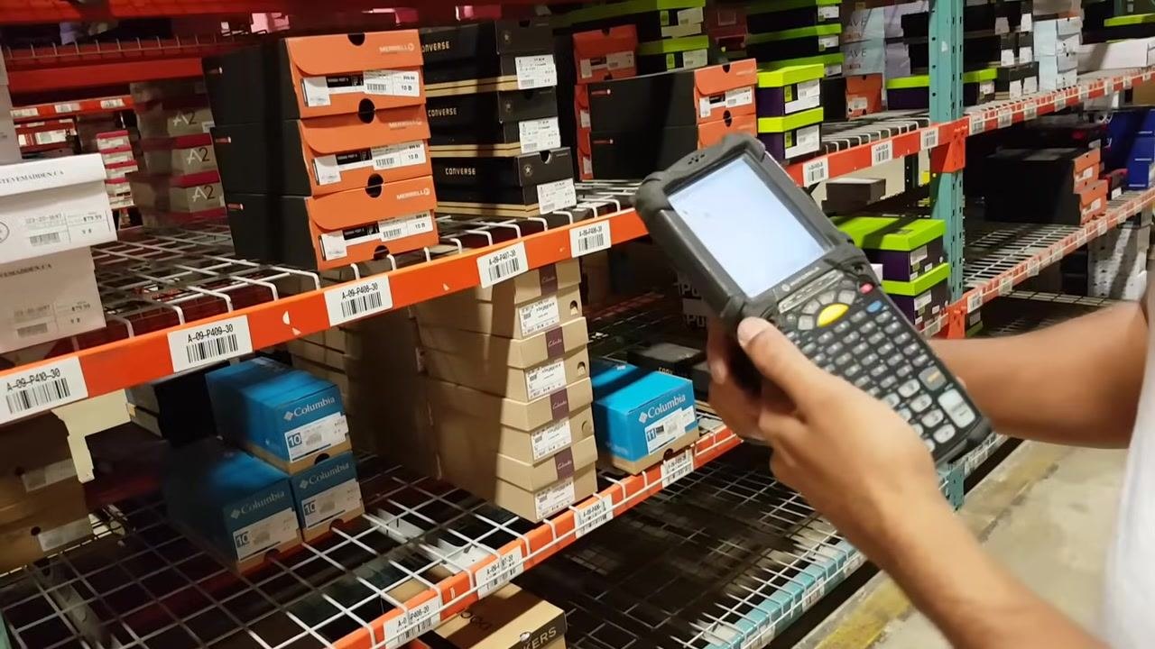 A worker stands in front of warehouse shelves filled with shoe boxes of various brands, holding an RF gun and comparing the device screen to the bin labels on the shelf. Bin labels and stacks of Columbia and Converse shoe boxes are visible.