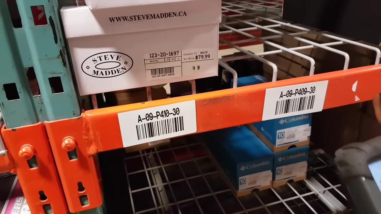A person points to a shelf label "A-09-P410-30" while holding a handheld scanner. Shoe boxes labeled "Steve Madden" and "Skechers Performance" are visible on the shelves.