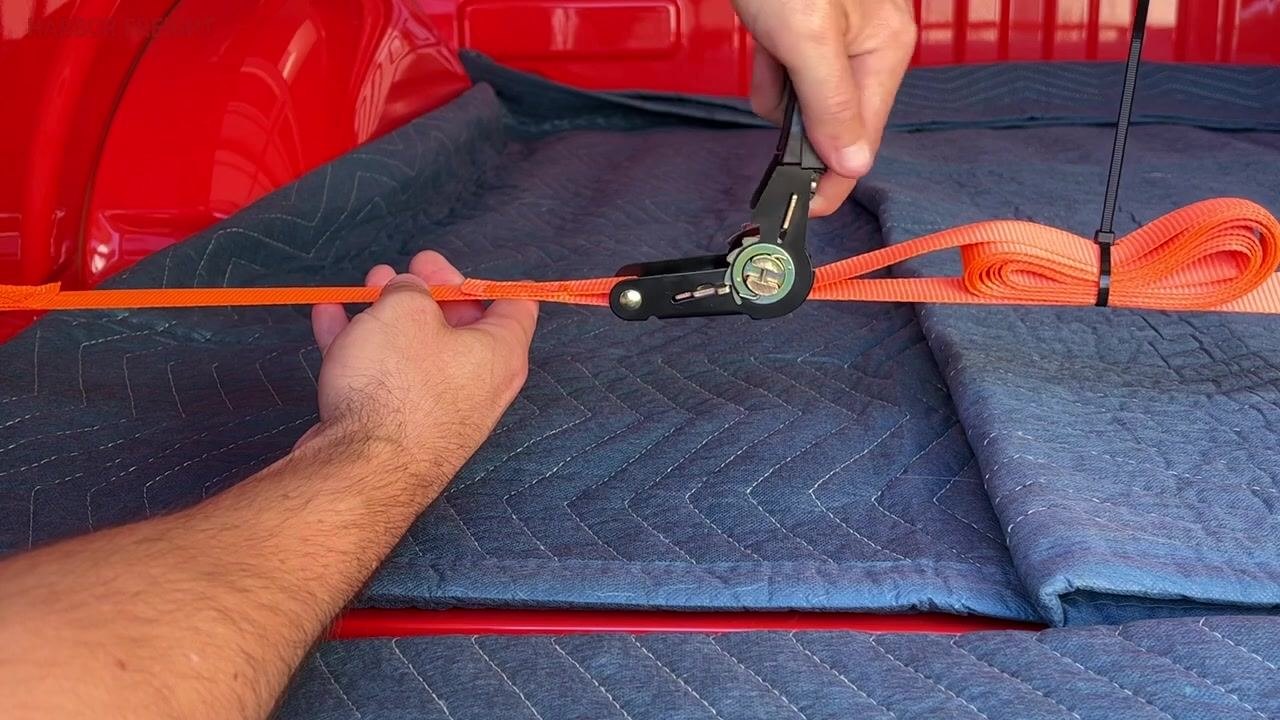 A hand holding the ratchet handle flat, with the orange strap being pulled free from the mechanism across the truck bed.