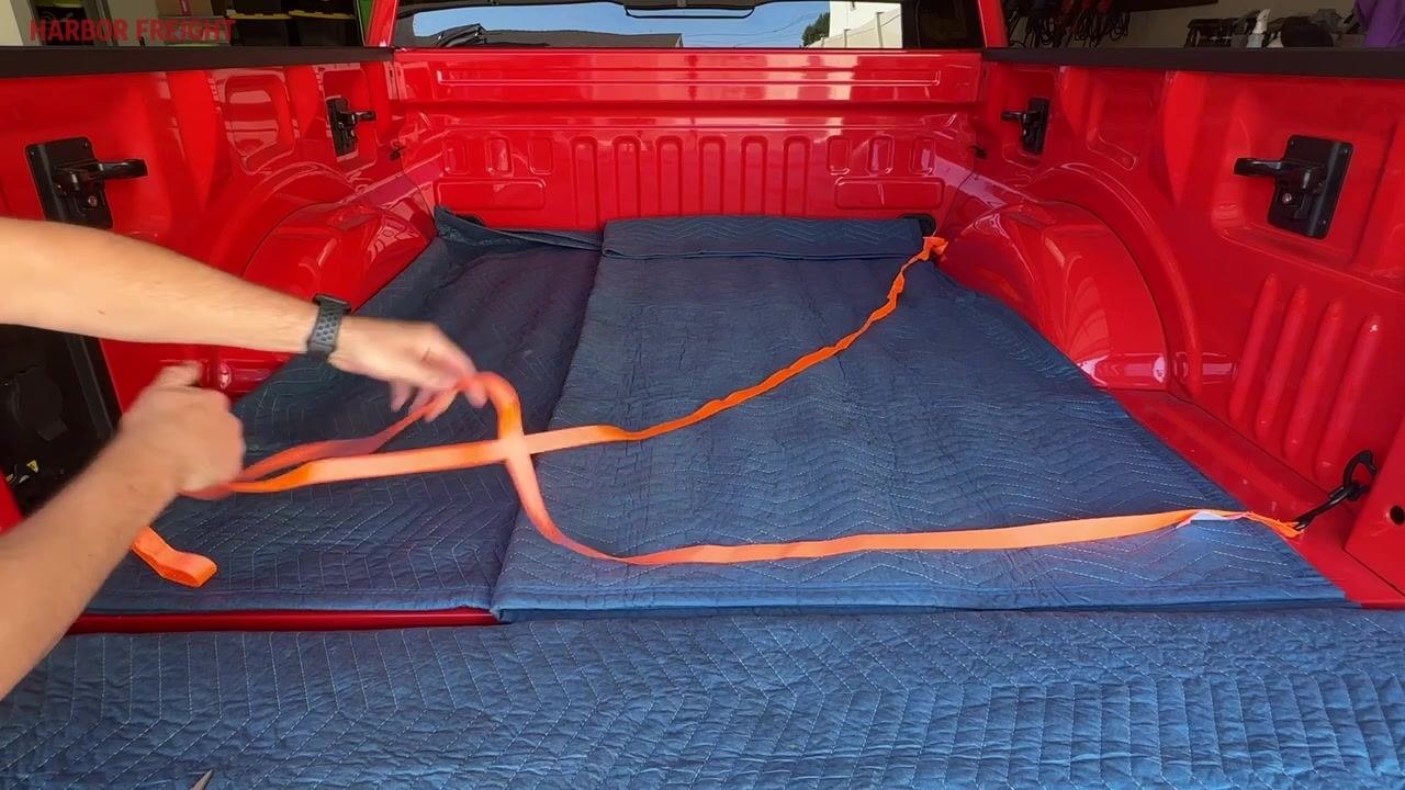 A hand pulling the orange ratchet strap free through the mechanism in the bed of a red truck lined with blue moving blankets, with the strap loose and no visible tension.