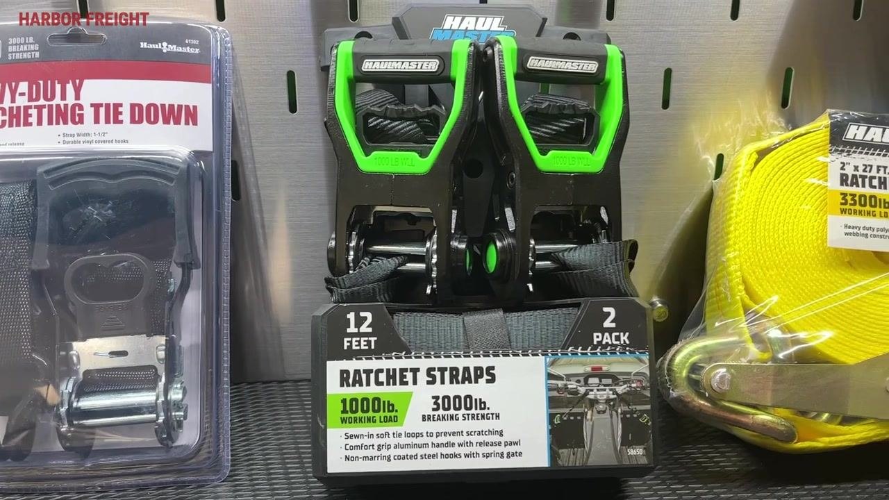 Retail display of green-handled ratchet straps rated at 1,000 lb. working load / 3,000 lb. breaking strength, alongside a yellow heavy-duty 2" x 27' strap rated at 3,300 lb. working load.