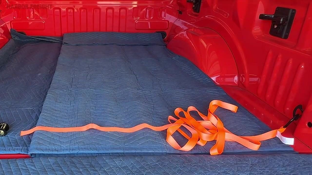 A hand holding the straightened orange strap in preparation for feeding it into the ratchet, with the rest of the strap laid out in the truck bed.