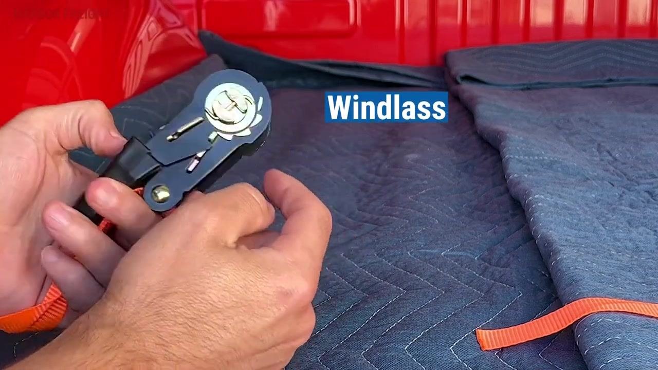 A close-up of hands holding a ratchet mechanism with the windlass labeled. The orange strap is visible, and the ratchet rests on a blue moving blanket in a red truck bed.