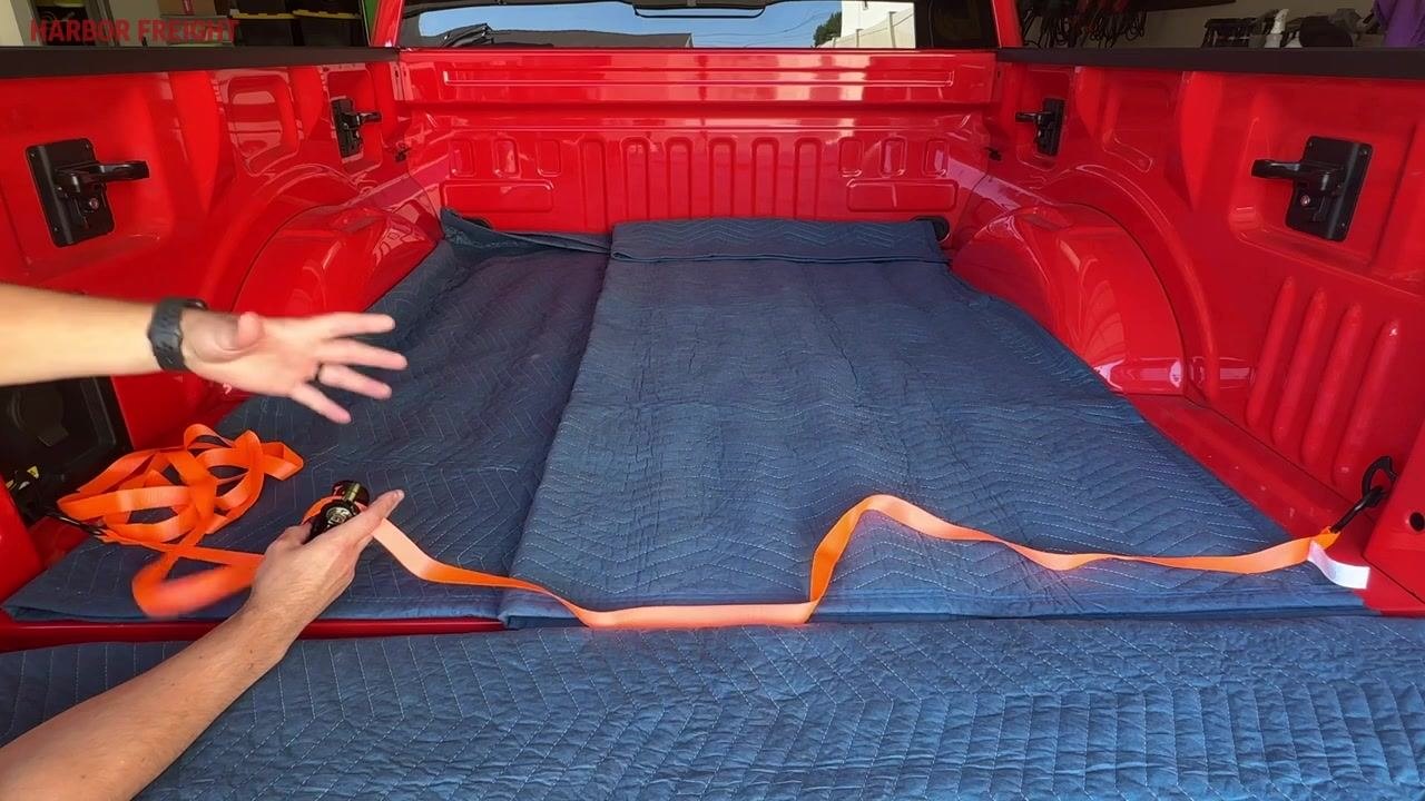 A hand positioning the ratchet mechanism upright on a blue moving blanket in a red truck bed, with the orange strap nearby.