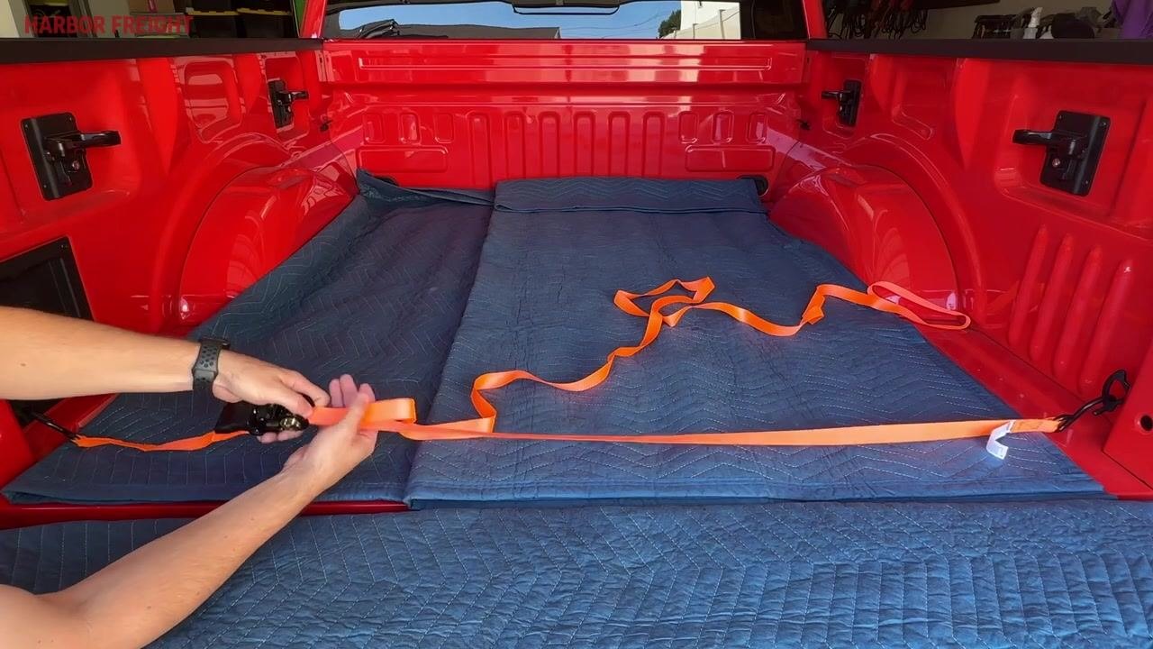A hand operating the ratchet handle, causing the orange strap to wind around the windlass, with some slack still visible across the truck bed.