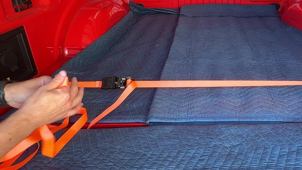 Hands holding a ratchet strap with excess orange webbing, preparing to manage the loose end in the bed of a red truck lined with blue moving blankets.