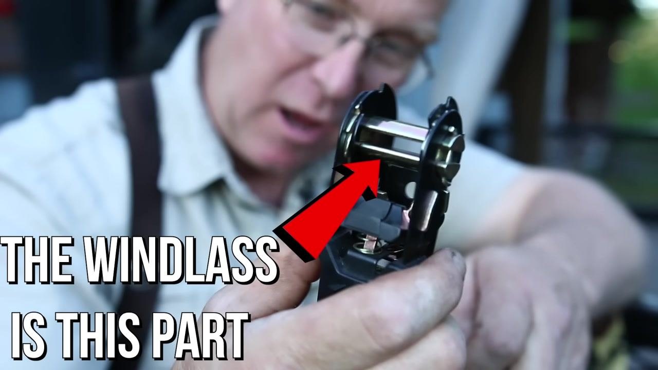 A person holds a ratchet mechanism in one hand and the detached strap in the other, standing beside a metal trailer outdoors.