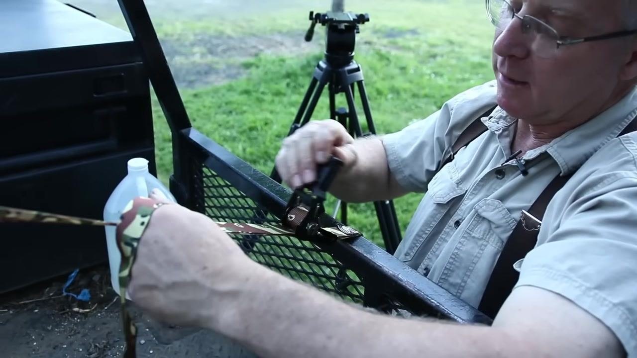 A person operates the ratchet mechanism with a camouflage strap threaded through it, attached to a metal trailer gate; a white plastic jug and tripod are visible in the background.