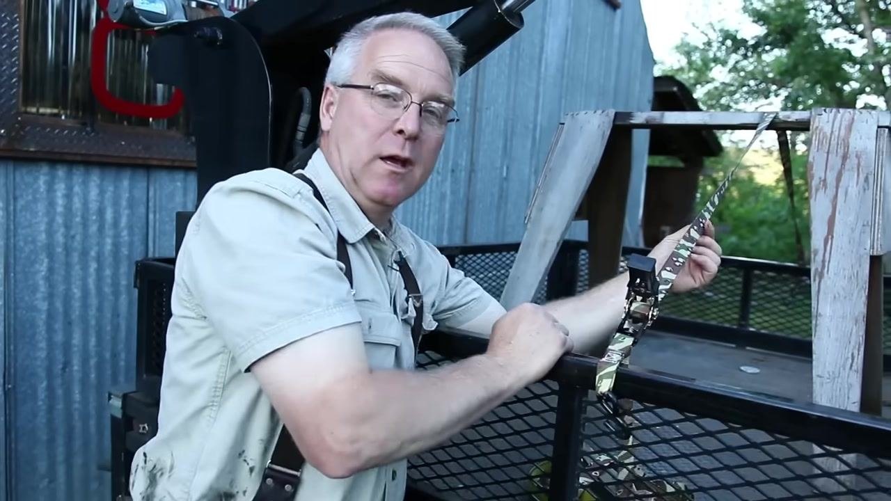 A person works to free a stuck ratchet strap by pressing the handle straight and pulling on the strap, with a tripod and jug visible in the background.