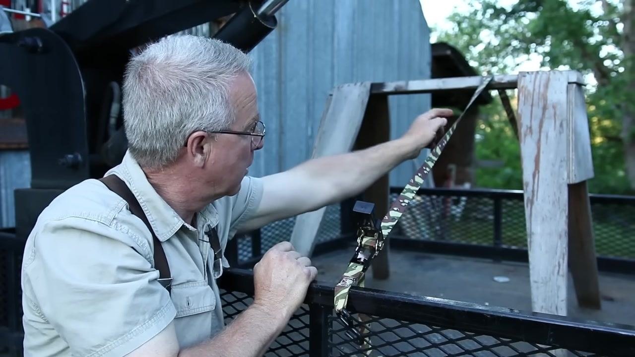 A person points to the ratchet strap to show the correct half-twist that prevents vibration, with a clear outdoor view of the strap's orientation.