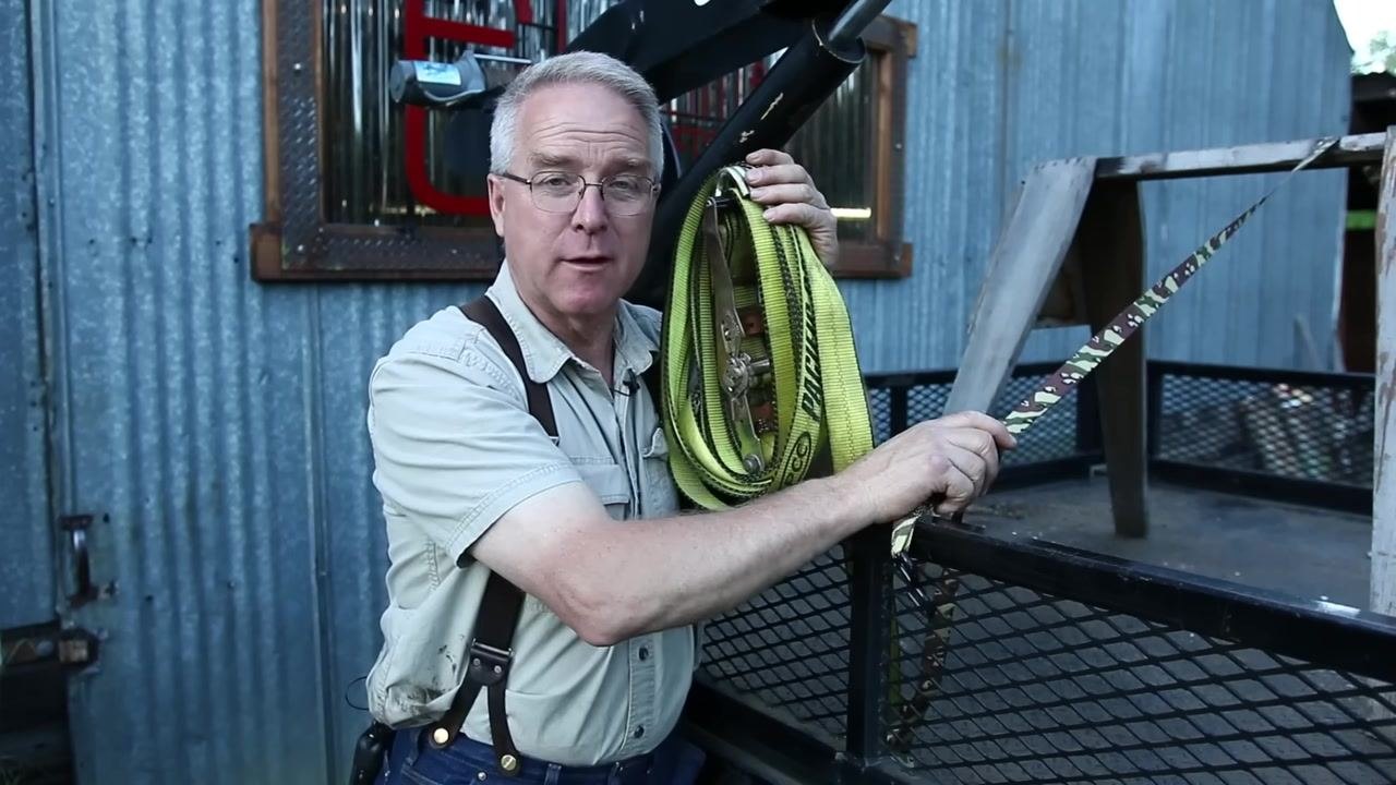A person stands next to a trailer, demonstrating a camouflage ratchet strap with a visible half twist spanning two points, in an outdoor setting with a wooden structure and metal fencing.